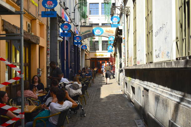 Brussels, Belgium - July 17, 2021: situated Loyalty corridor, Jeanneke Pis, less famous than Manneken Pis, fountain statue erected in 1987, female version of Manneken Pis, Grand Place. Narrow street at the end on the right side statue placed in  a carved wall terraced house. African waiter is taking an order.