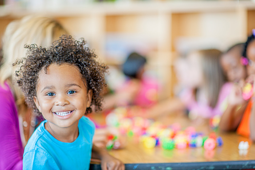 A diverse group of children sit around a desk in preschool. An adorable kid smiles at the camera.