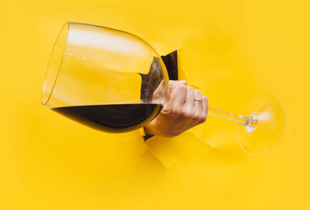 A right woman's hand emerges through a torn hole in yellow paper with a large glass of red wine. The concept of alcoholism, drunkenness and hangover. stock photo