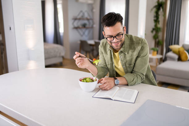 istock Young man eating a salad while reading a book at home. 1329420767