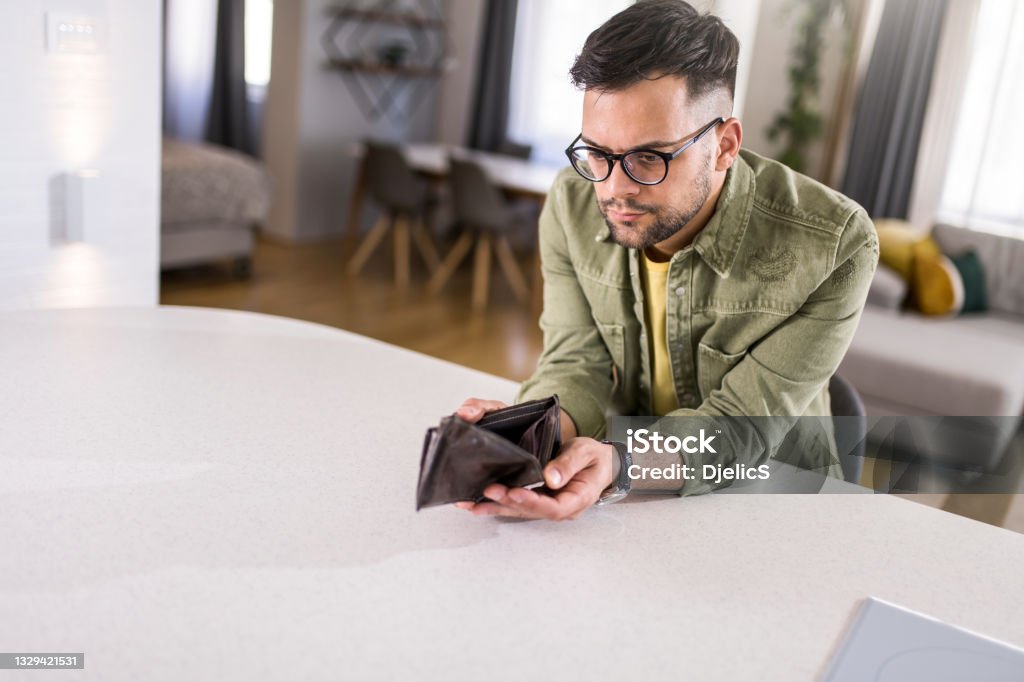 Worried young man having financial problems. Young man sitting at home with an empty wallet. He is left without money and thinking what to do next. Empty Wallet Stock Photo