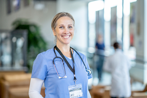 A nurse wearing blue scrubs smiles confidently at the camera. She is alone, but there are doctors and nurses walking around in the clinical space behind her.