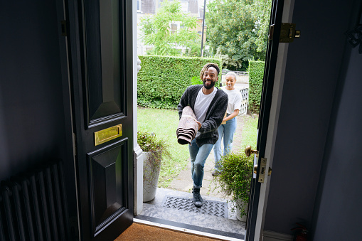 Interior viewpoint of man and woman in their 30s wearing casual clothing and approaching front door carrying rolled-up rug and cardboard box.