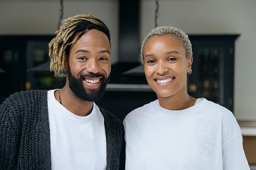 Portrait of bearded man and short haired woman wearing casual clothing and standing in kitchen smiling at camera.