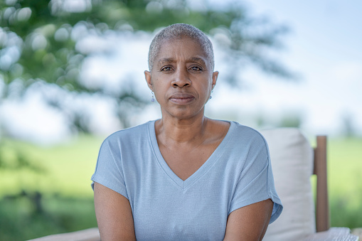 An African American woman with short grey hair sits outside in the summertime and has a reflective, pensive look on her face. Her hair loss signifies a history of chemotherapy treatment.