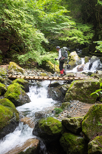 A senior man hiking a mountain trail crossing a wood bridge over a freshwater mountain stream waterfall.