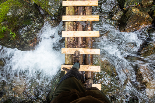 A personal perspective view looking straight down at feet crossing a wood hand made bridge crossing a mountain stream below.