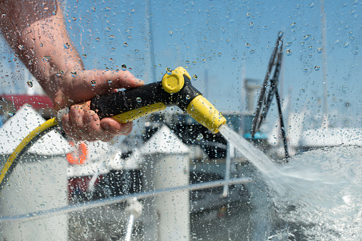 Hand holding water spray gun. Man washing boat with pressure water system. View from inside boat. Focus on water drops. Yacht maintenance concept.