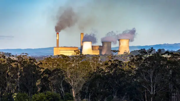 Brown coal-fired power station amongst native trees and hills, emitting smoke from twin smokestacks and heat from three cooling towers: Yallourn Power Station in Latrobe Valley, Victoria to be closed in 2028 and be replaced by a huge battery: climate change, global warming, harmful to health, environmental issues.