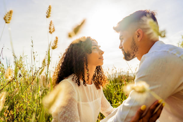 scena romantica di giovane coppia appassionata multirazziale innamorata che si guarda negli occhi tra un'alta vegetazione erbosa al tramonto o all'alba con effetto retroilluminato dal sole sogno romantico girato dagli amanti nella natura - heterosexual couple couple sensuality young couple foto e immagini stock