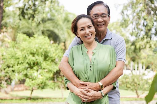 Positive elderly couple standing in park, husband hugging wife from behind and looking at camera