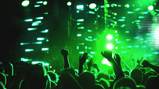 Rear view of large group of people enjoying a concert performance. There are many hands applauding and taping the show. Green lasers and spot lights firing from the stage.
