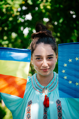 Portrait of a proud Romanian teenage girl holding a Romanian flag and the flag of the European Union (EU). She is wearing the traditional national clothing known as 'Ie'. She is happy and looking at the camera and smiling.