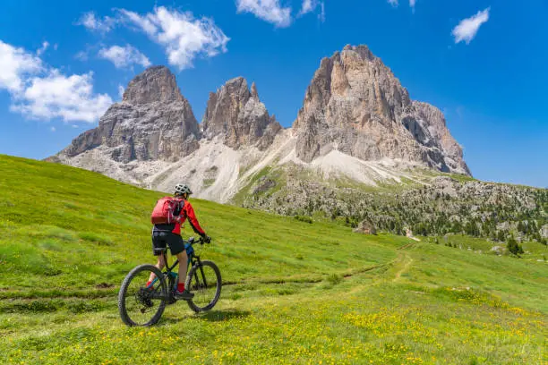 pretty beautiful senior woman riding her electric mountain bike below famous Sassolungo summit in Sella Dolomites of Selva Wolkenstein, Val Gardena, South Tirol and Trentino, Italy