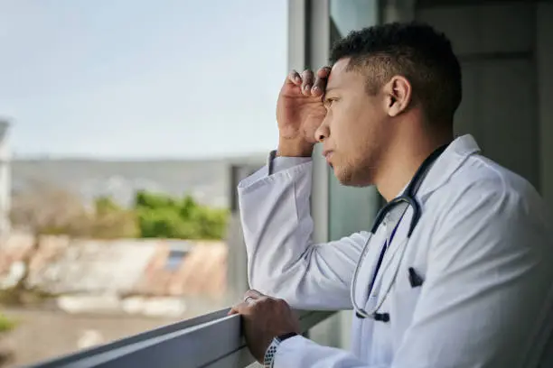 Young male doctor wearing a lab coat deep in thought while looking out of a hospital window l