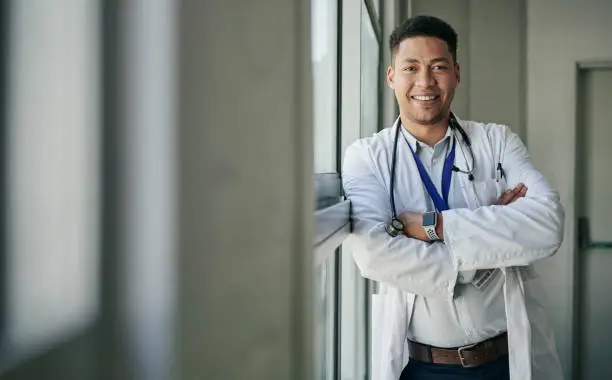Portrait of a smiling young male doctor wearing a lab coat leaning on a window in the corridor of a hospital