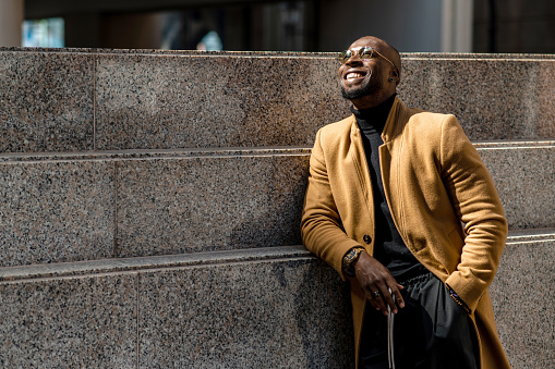 Portrait of attractive black man in stylish clothing leaning on a city wall looking up with smiley positive face.