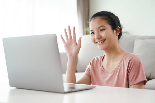 istock Young Asian woman using laptop computer for online video conference call waving hand making hello gesture in the living room. 1329485815
