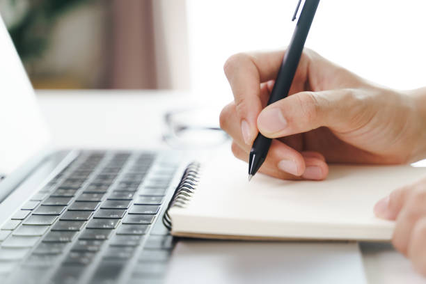 istock Close up of young man in casual cloth hands writing down on the notepad, notebook using ballpoint pen with laptop computer on the table. 1329485841