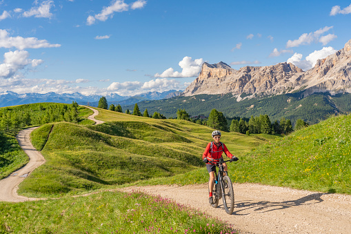 nice and active senior woman riding her electric mountain bike on the Pralongia Plateau in the Alta Badia Dolomites with awesome Sasso die Santa Cruce summit in Backg, South Tirol and Trentino, Italy