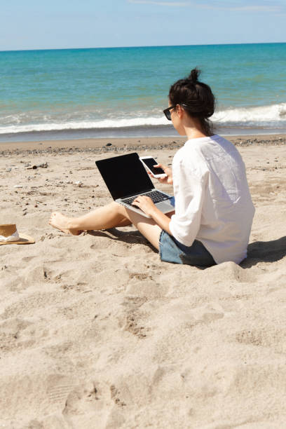 istock Young woman using laptop computer and holding a cell phone on a beach. Vacation lifestyle communication. Freelance work concept 1329488122
