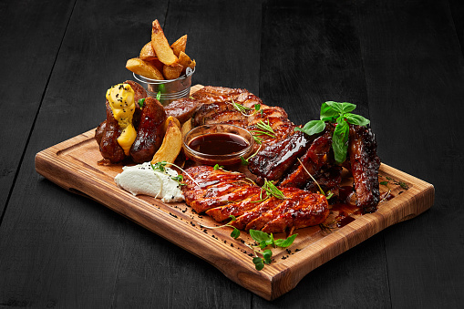 Delicious grilled assorted meat platter for company. Spicy sausages, lamb ribs, sliced pork steak and chicken fillet served on wooden board with crispy potato wedges and sauces garnished with greens