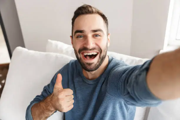 Happy young man sitting on a couch at home, taking a selfie, thumbs up