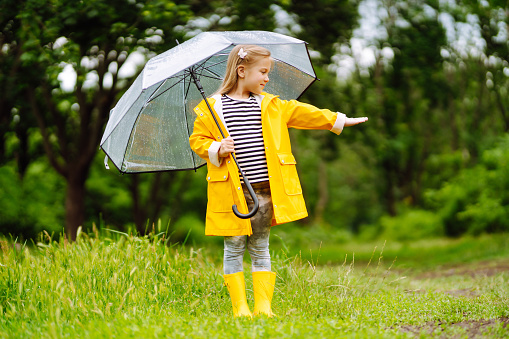 Funny girl with transparent umbrella enjoys the rain. She is wearing  yellow raincoat and rubber boots. Little girl plays outdoors in nature. Happy childhood concept.