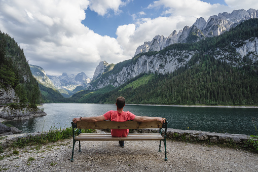 Man relaxing on bench in front of Dachstein Mountains reflected in Gosau lake, Austria.