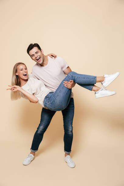 Full length portrait of a cheerful young couple Full length portrait of a cheerful young couple standing together over beige background, man holding girlfriend on his arms couple isolated wife husband stock pictures, royalty-free photos & images