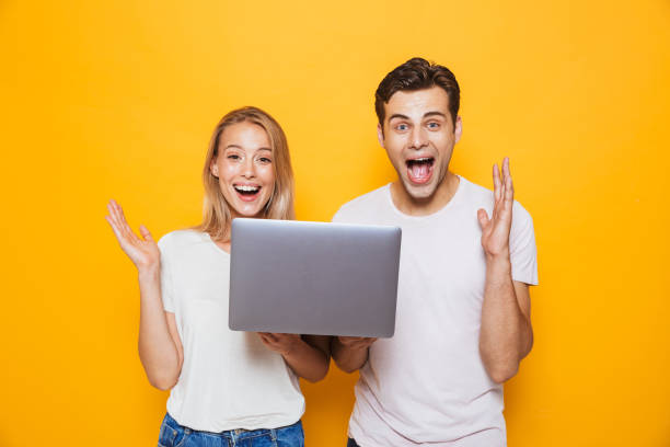 heureux excité jeune couple amoureux isolé sur fond de mur jaune à l’aide d’un ordinateur portable. - women computer couple surprise photos et images de collection