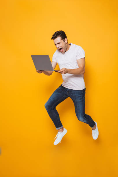 istock Young man jumping isolated over yellow wall background using laptop computer. 1329503488