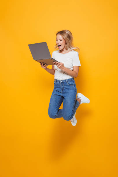 giovane donna eccitata che salta isolata sullo sfondo della parete gialla usando il computer portatile. - saltare foto e immagini stock