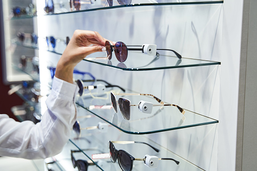 Close up of young woman standing by glass showcase and choosing shades in eyeglasses store