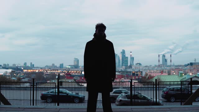 The camera zooms in on a guy who is standing against the background of a big city. Cool gloomy silhouette. In the background, high-rise buildings and pipes