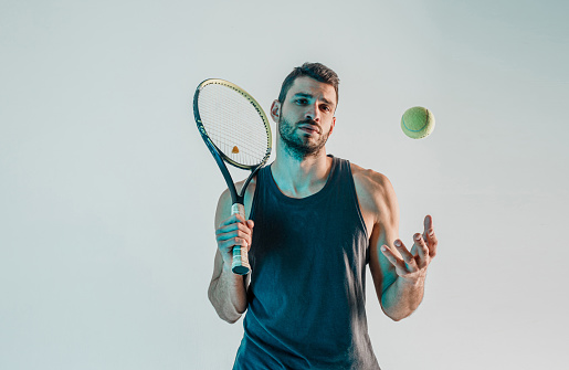 Serious tennis player hold racquet and throw up ball. Front view of young bearded european sportsman looking at camera. Isolated on turquoise background. Studio shoot. Copy space