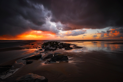 A beach on a stormy night during sunset. The water is moving quickly and there are rocks going into the water. The sky is filled with dark clouds, the sun is shining on them in the far background. The water reflects this phenomenon.