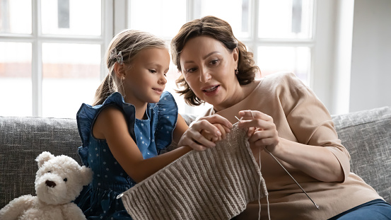 Caring old Caucasian grandmother teach small teen granddaughter knit with needles relax together at home. Loving mature granny and little grandchild have fun rest in living room with hobby activity.