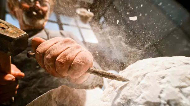 Close-up of stonemason's hands chiselling surface of a white stone sculpture in the workshop.