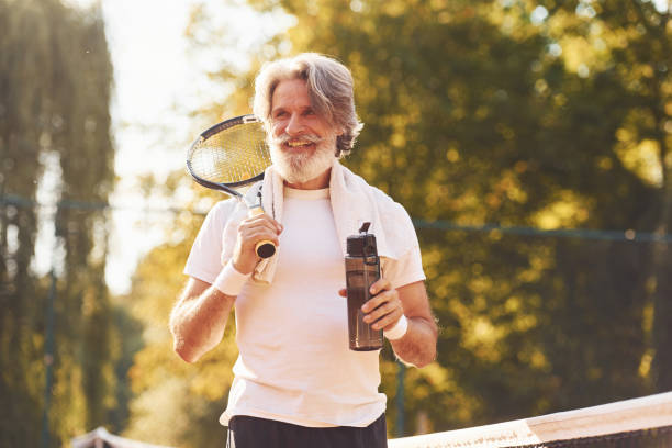 Taking break and drinking water. Senior stylish man in white shirt and black sportive shorts on tennis court Taking break and drinking water. Senior stylish man in white shirt and black sportive shorts on tennis court. tennis senior adult adult mature adult stock pictures, royalty-free photos & images