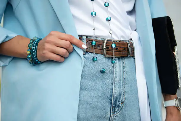 Detail of the body of a model dressed in light blue high-waisted jeans, an ornamented leather belt and a blue jacket. Jewelry from blue stone beads and bracelet.