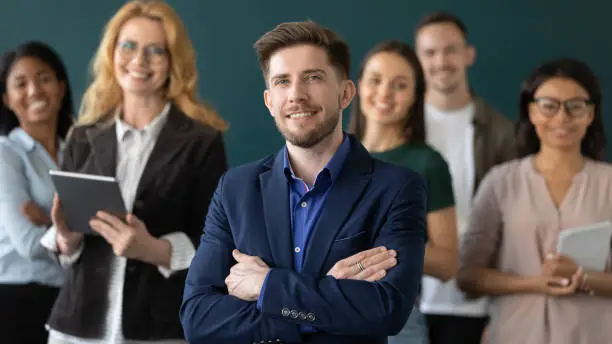 Happy confident male business leader posing with team in blurred background. Young businessman, director, manager with group of office employees looking and smiling at camera. Head shot portrait