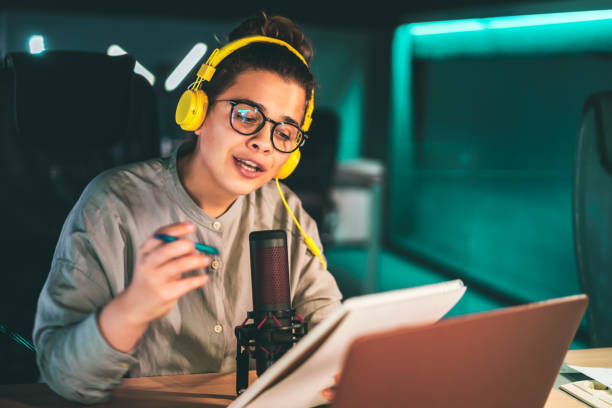 Woman recording an audio podcast in modern studio Young woman recording an audio podcast in a modern studio, wearing headphones and hipster glasses, speaking to the microphone and holding a notepad media interview photos stock pictures, royalty-free photos & images