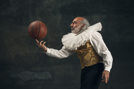 One old man having fun, playing with basketball ball isolated on dark vintage background. Retro style, comparison of eras concept. Elderly male model like sportsman