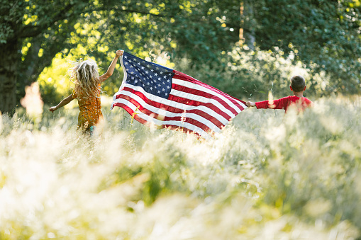 A patriotic scene with two children holding a United States flag in a sunlit grassy field.  A concept of freedom or celebrating a holiday like the 4th of July, Independence Day.