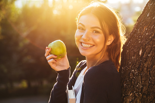 Beautiful young woman holding apple while standing at the tree in the park