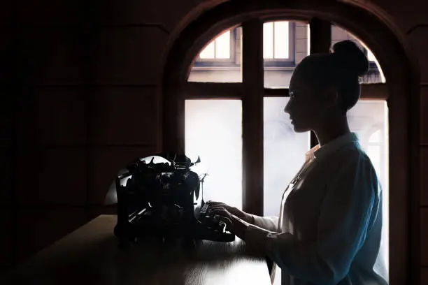 young beautiful woman author at a typewriter, writes a text, inspiration in creative work. Retro vintage typewriter side view, silhouette in a dark room against the background of a window