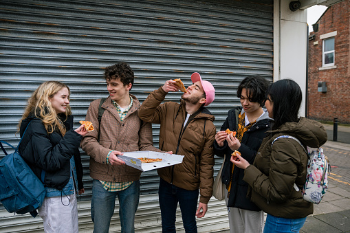 A front-view shot of a group of female and male teenage friends standing against a shutter on a street in Wallsend, North East England. They are all sharing and eating fish and chips out of paper wrappings.