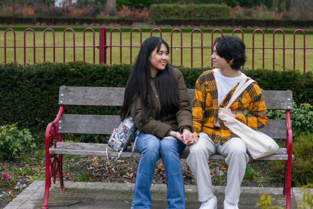 I Love Spending Time with You! A front-view shot of a young teenage couple sitting on a wooden bench in a public park in Wallsend, North East England. They are looking at each other and holding hands. teen romance stock pictures, royalty-free photos & images