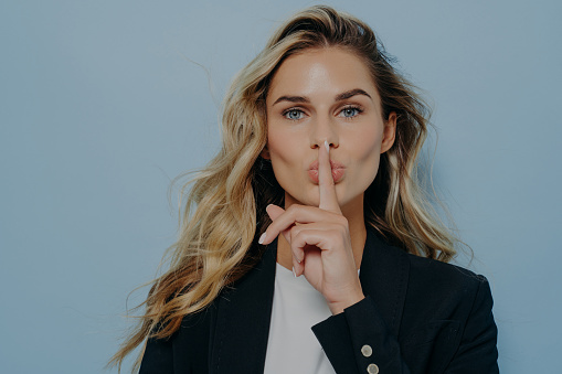 Blonde beautiful young woman wearing black coat making shush gesture with her hand, telling someone to be quiet, dont make any sound, standing isolated next to blue wall. Be silent concept